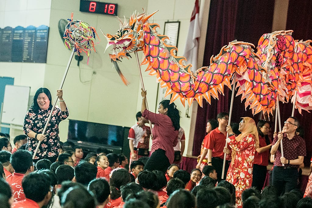 People perform a colorful dragon dance with orange dragon props on poles before a large seated crowd indoors.
